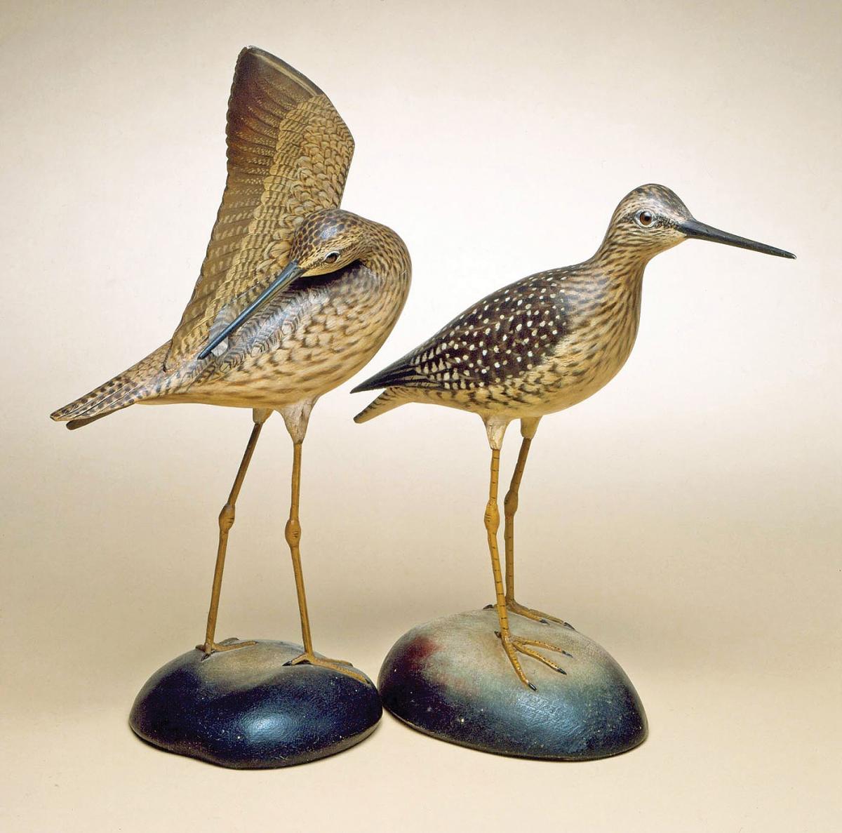 A pair of yellowlegs birds, carved in wood, one standing and the other reaching its beak under its wing