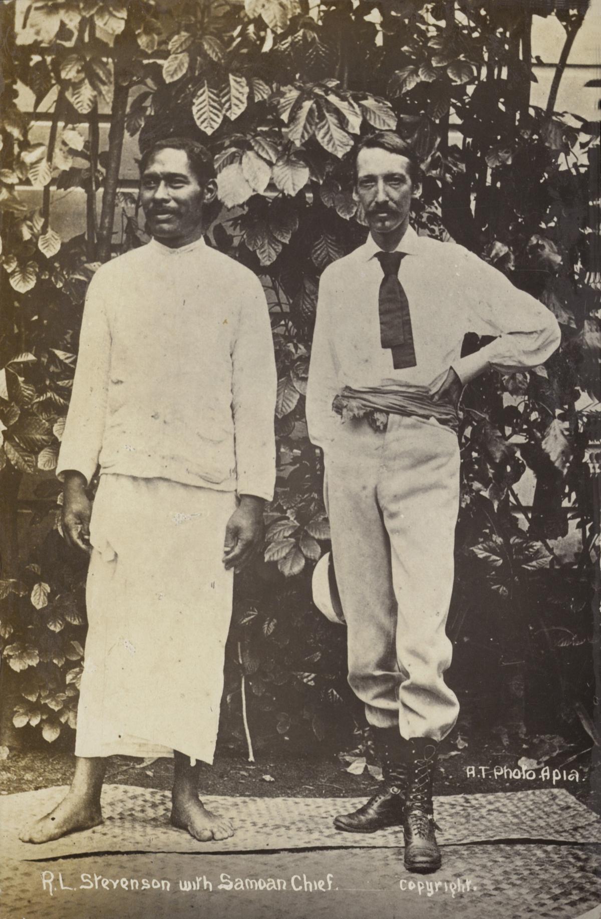 Black and white photo of Stevenson in a white shirt and hand on his hip, standing next to a Samoan chief, who is dressed in traditional white Samoan clothing