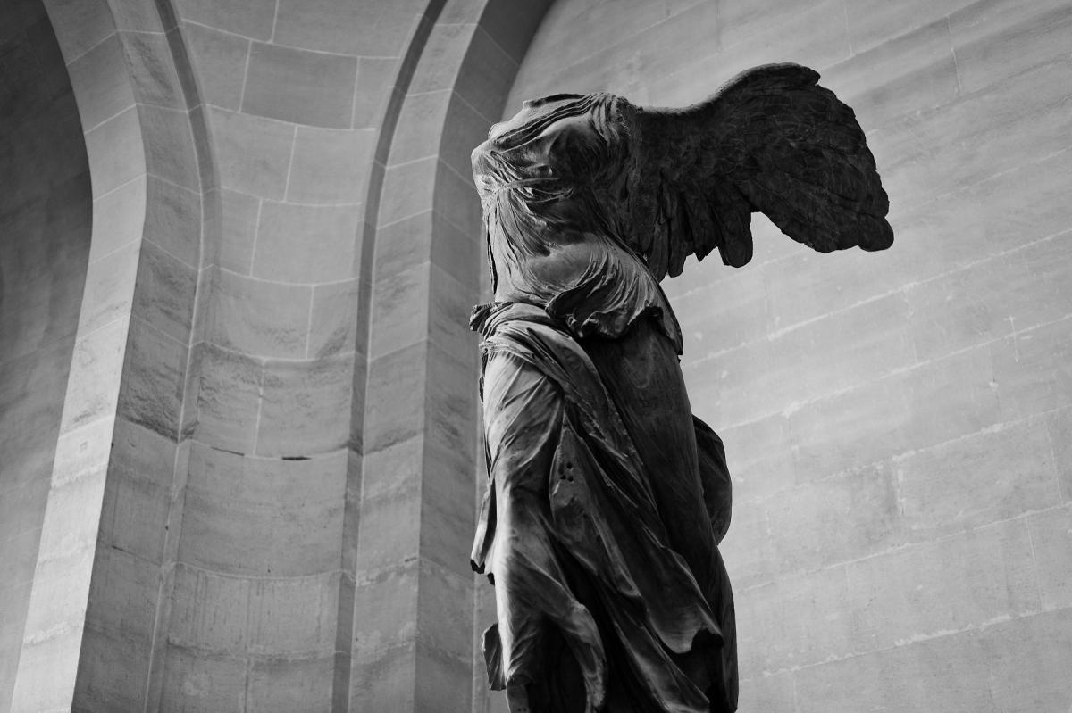 Black and white photo of a winged statue with no head, viewed from afar.