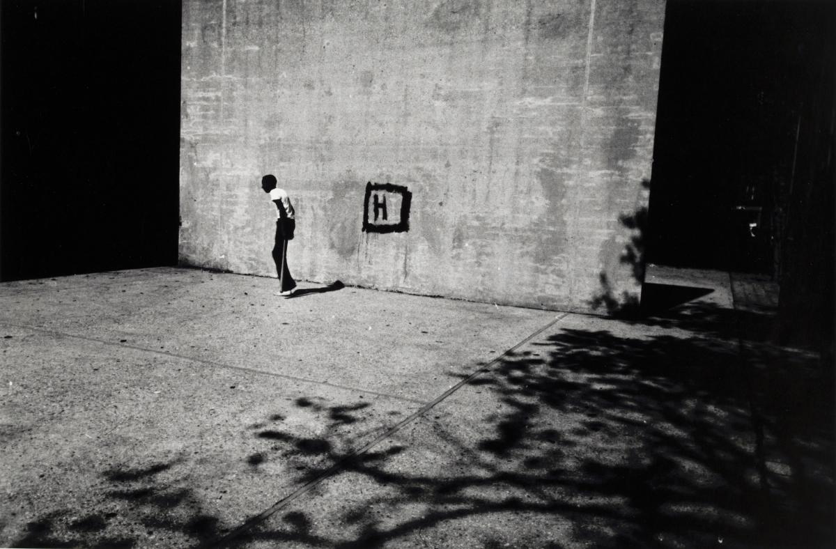 A thin boy holding a stickball bat in a handball court, the wall marked with a square and the letter H, as dark shadows bracket the image