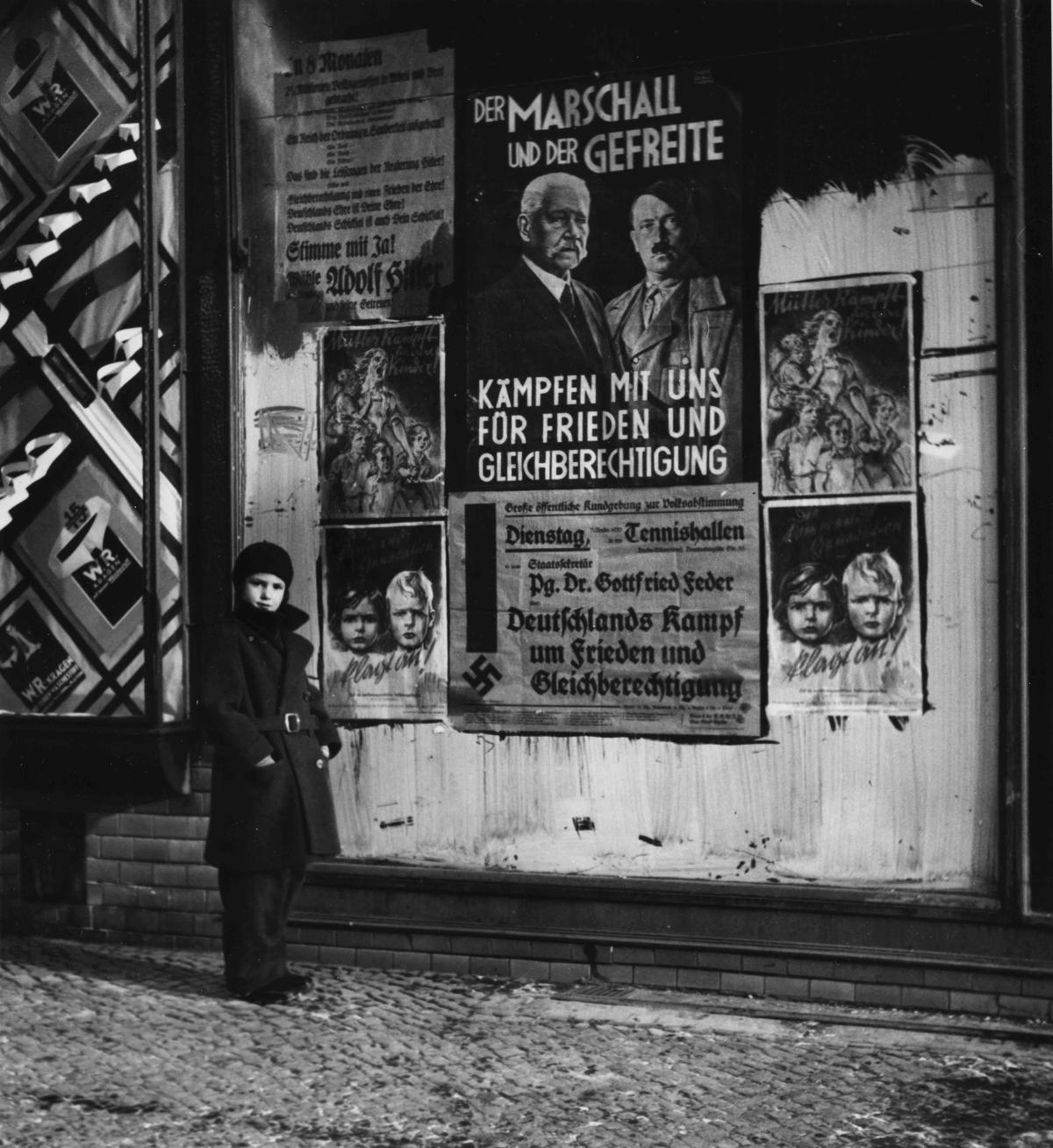 black and white photo from 1930s with small girl in front of Nazi poster
