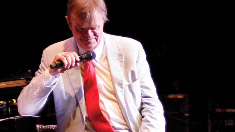 A portrait of Garrison Keillor in a white coat and khaki pants, holding a microphone.