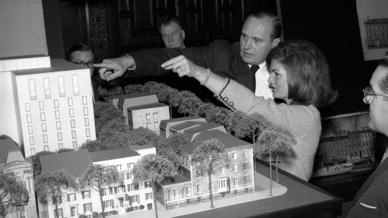 Black and white photo of Jacqueline Kennedy gesturing to an architectural model with a man 