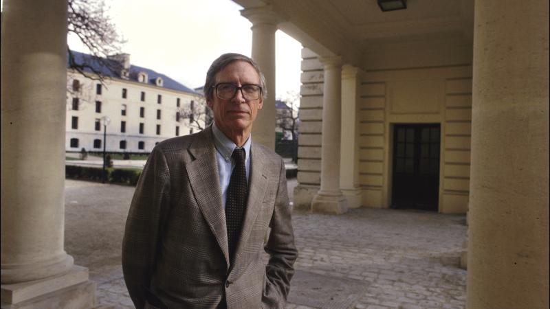 A portrait photo of John Rawls standing amidst neoclassical columns.