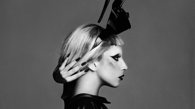 Side profile of Lady Gaga, wearing a black sculptural headdress, reaching her hand around the back of her neck