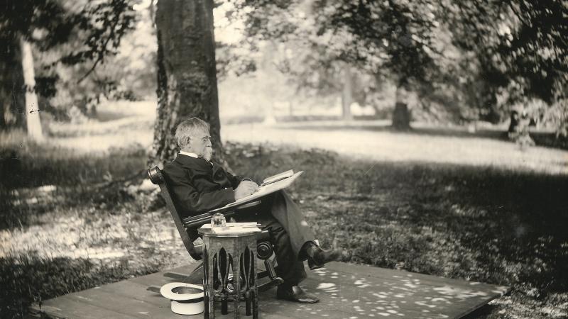 Wallace sits in a rocking chair, writing in pen on a notebook, legs crossed