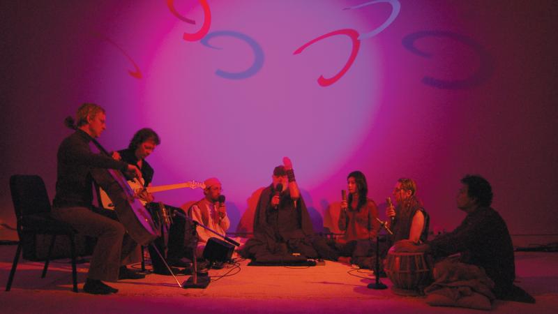Seven performers sit in a darkened room, holding instruments, lit only by a nimbus of purple light