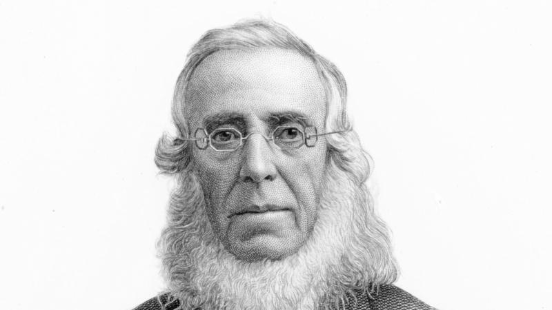 Cooper wearing octagonal eyeglasses, with a white beard no mustache, in a suit coat and bowtie