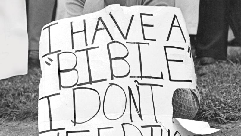 Black and white photo of a woman sitting on a curb holding a sign of protest with her chin on her fist.
