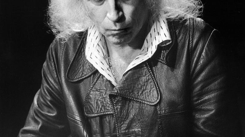 Black and white photo of an aged musician, slightly balded, with long, white, frizzy hair. He wears a leather jacket and collared shirt underneath.