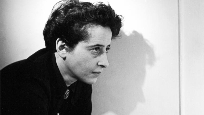 Arendt, with short dark hair, holding a cigarette, leaning on a countertop and facing to the right