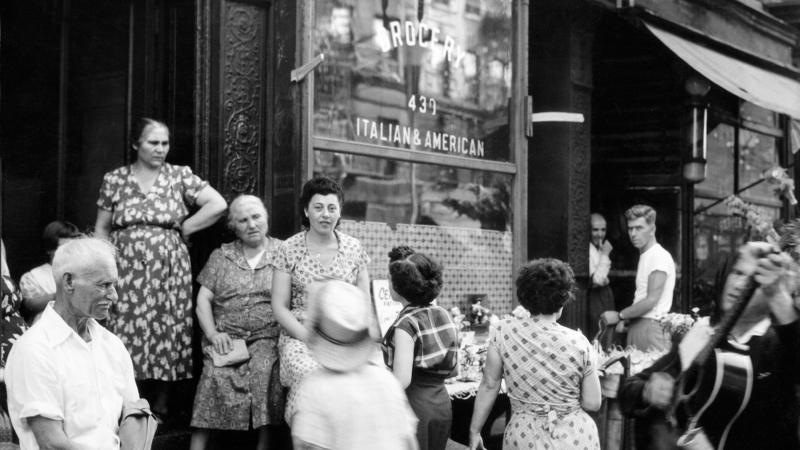 Black and white photo of Italian immigrants in Little Italy congregating outside a storefront. 