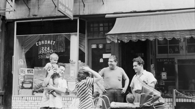 Black and white photo of Italian immigrants standing outside a storefront. One man is holding a baby up in the air, while a woman and two other men stand around a baby stroller.