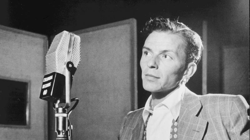 Black and white photo of Frank Sinatra standing before a microphone on stage.