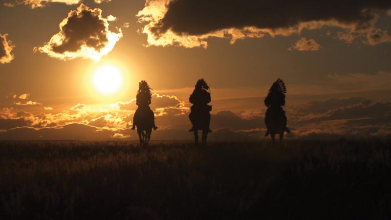 Color photo of three men on horseback riding into the glimmering sunset.