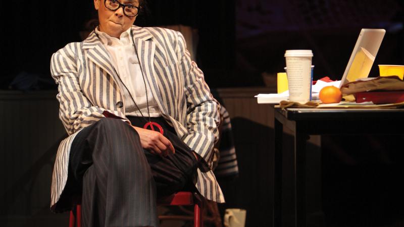 Color photo of Anna Deavere Smith playing a character on stage, sitting in a chair with her legs crossed.