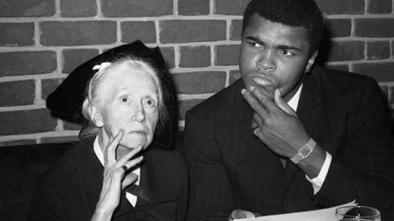 Black and white photo of poet Marianne Moore seated next to Mohammed Ali, who stares, intrigued, fingers on his chin, toward something else in the room.