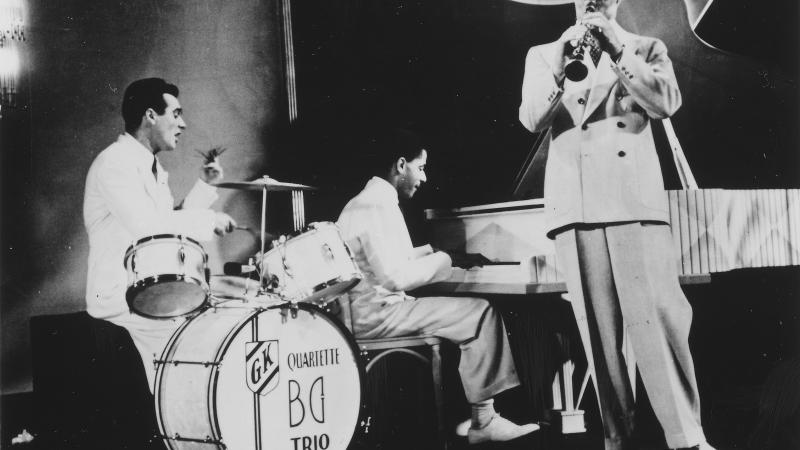 Black and white photo of a jazz trio, with one man on the drums, another on the piano, and the last one on the clarinet.