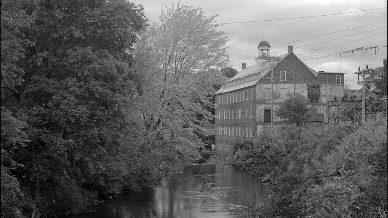 black and white photograph of a stream, building off of the bank