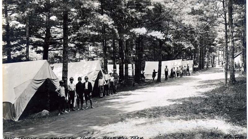 black and white photo of a row of tents, boys standing in front of them