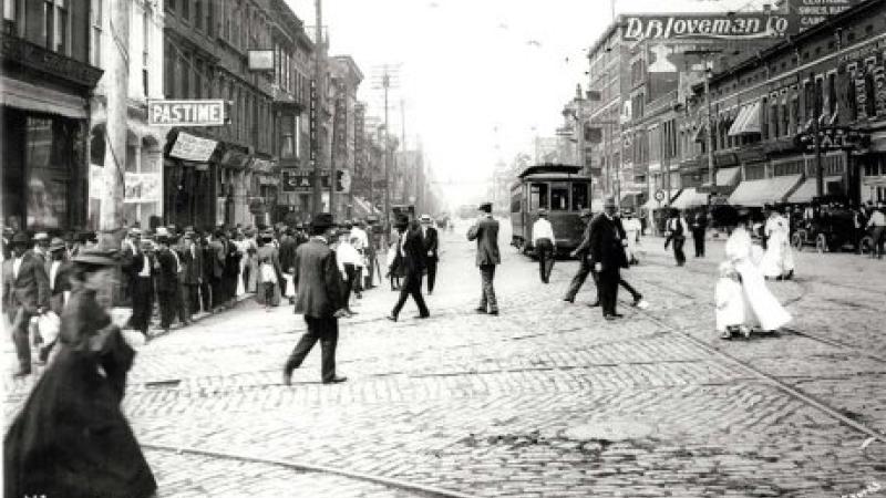 Black and white photo of a busy city street with people crossing and a trolley in the distance.