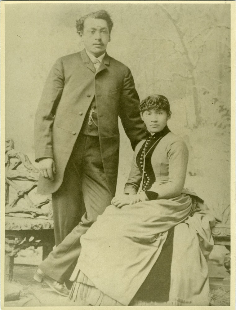 Mingo stands, with his hand on his seated wife's shoulder, both dressed in their wedding clothes