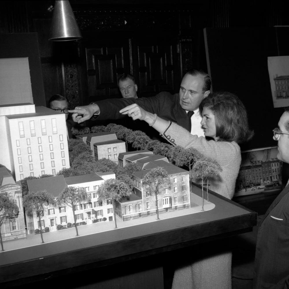 Black and white photo of Jacqueline Kennedy gesturing to an architectural model with a man 