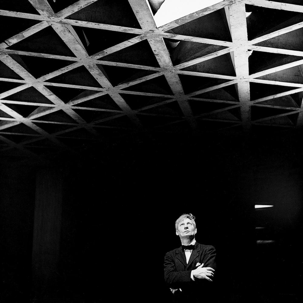 Black and white photo of Louis Kahn standing in a dark room, looking at a decorative ceiling. 
