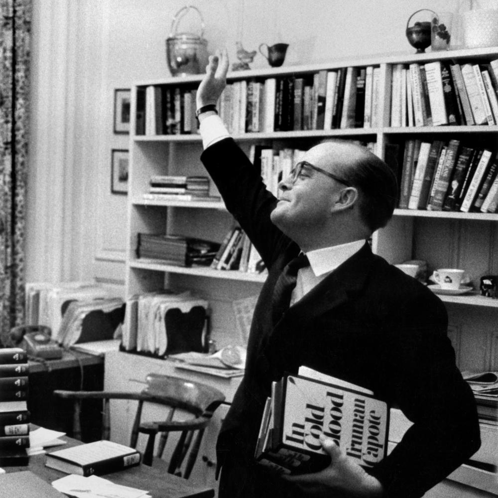 A black and white photo of Truman Capote waving his hand in the air in front of a bookshelf.
