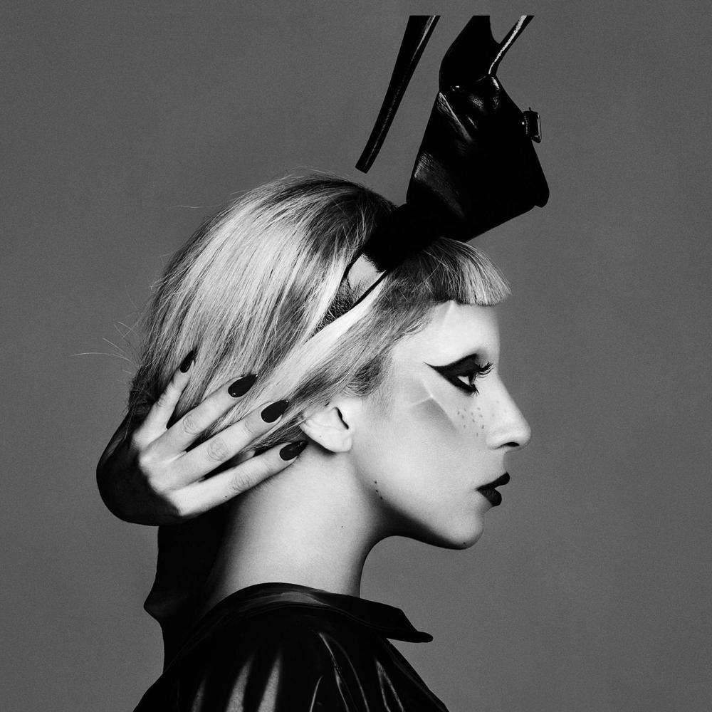 Side profile of Lady Gaga, wearing a black sculptural headdress, reaching her hand around the back of her neck