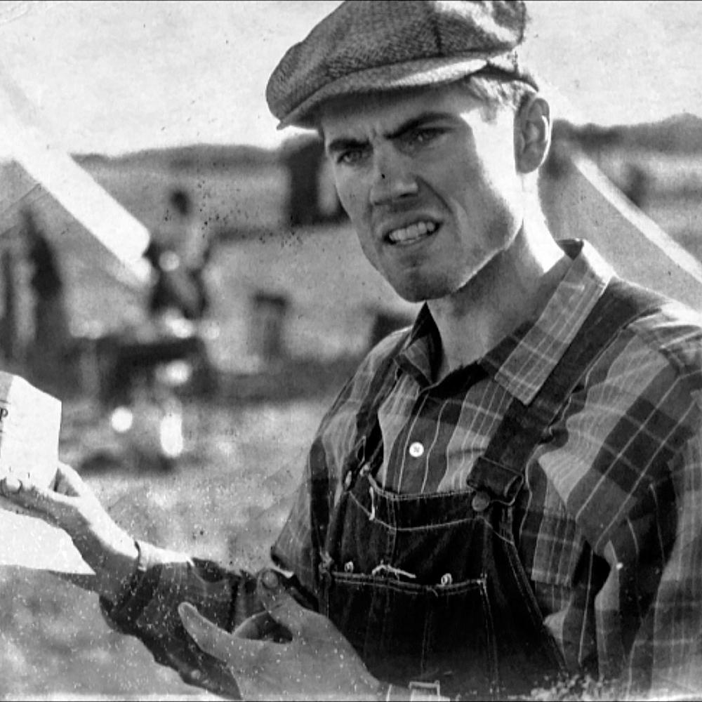 black and white film still of a man holding up a paper, wearing overalls and a hat
