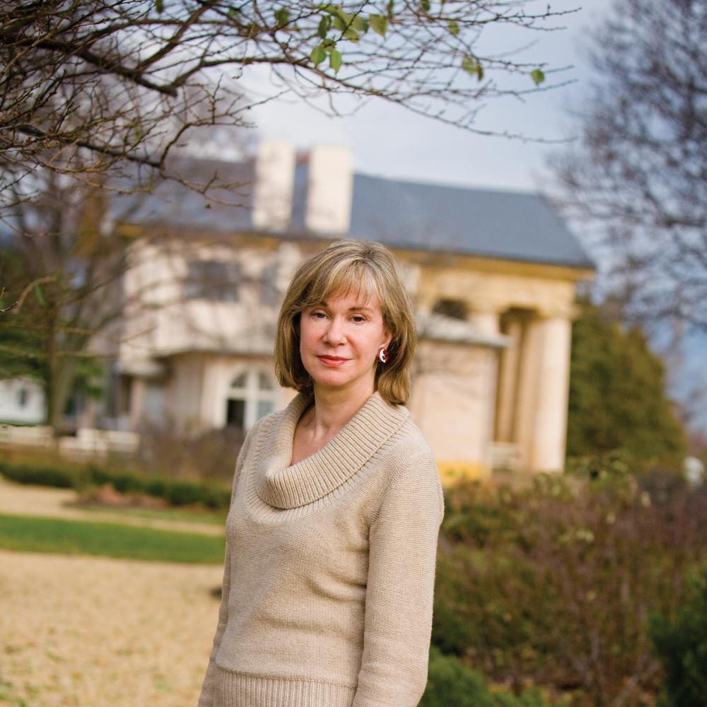 Brown Pryor in a tan cowl neck sweater, standing in a yard with a white house in the background