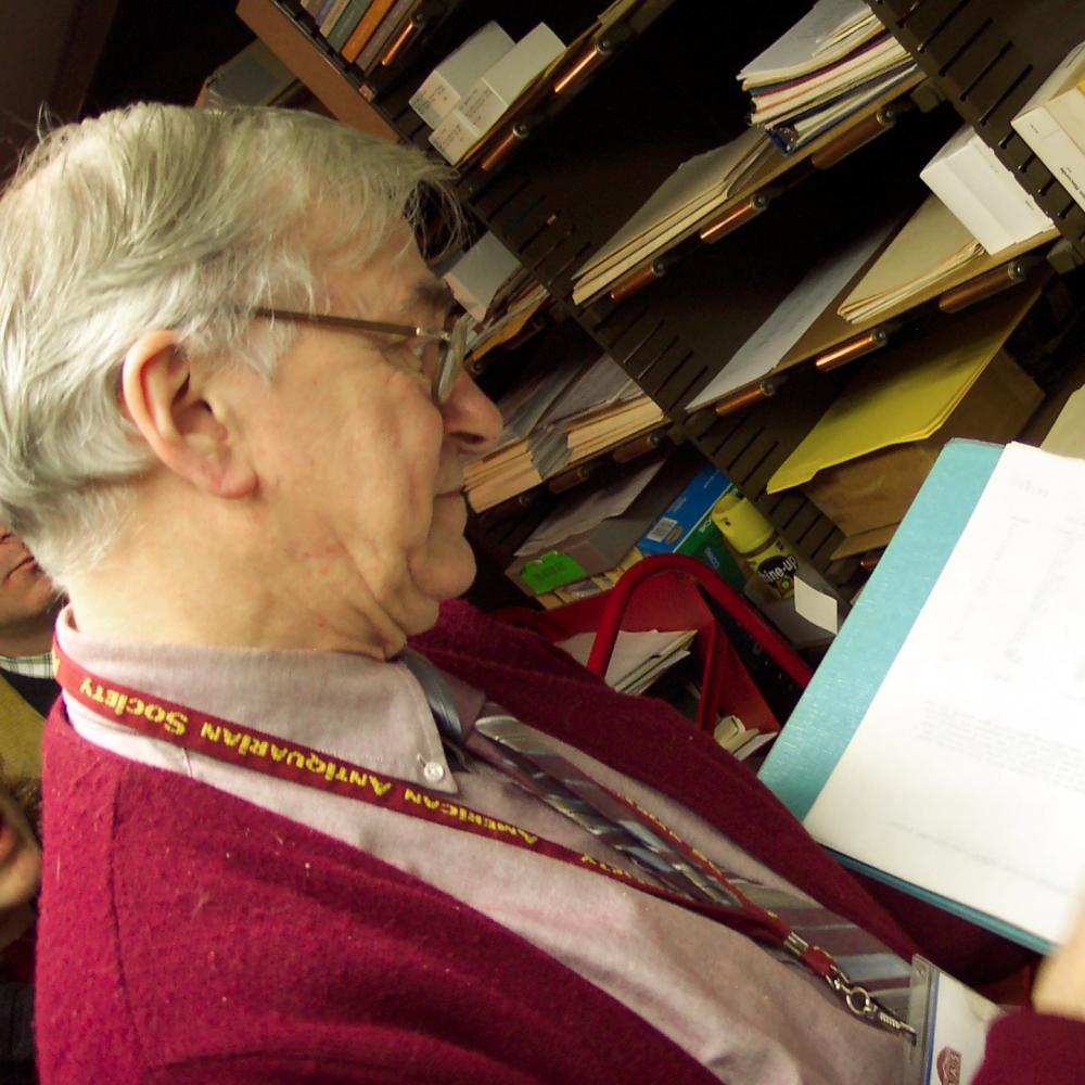 Lampi, wearing a red sweater, reads and holds an open binder in the library stacks