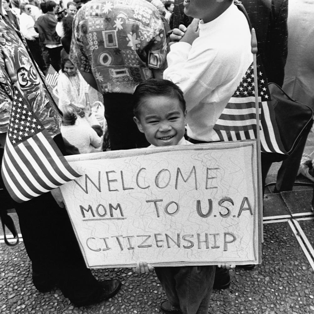 A young boy holds up a sign, flanked by American flags, that reads "Welcome Mom to USA citizenship"