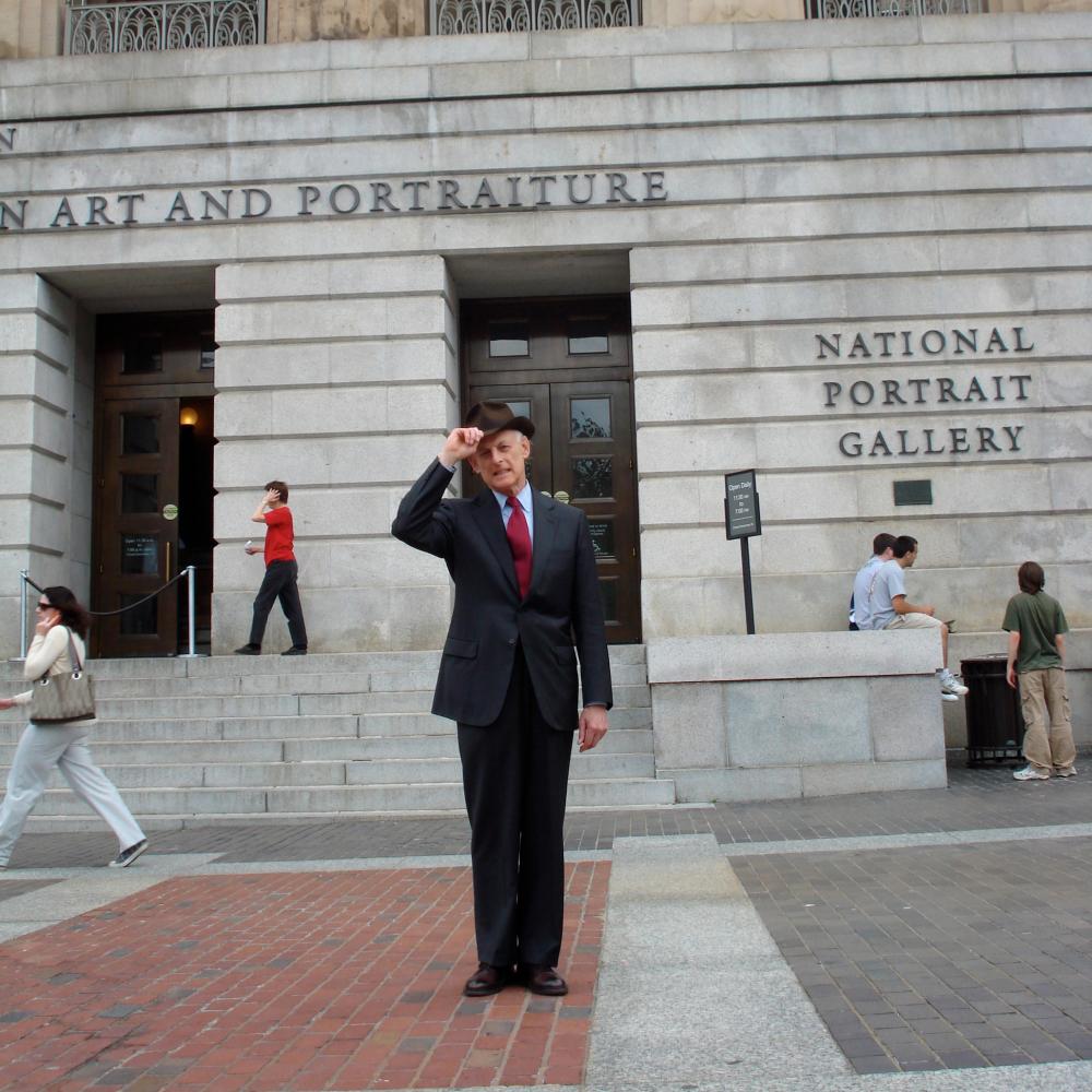 Photograph of a man in a suit touching brim of hat, outside of National Portrait Gallery