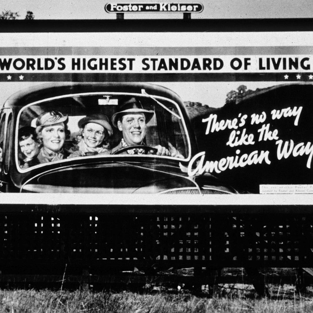 black and white photograph of a billboard showing a smiling family in a car, text reads "there's no way like the American way"