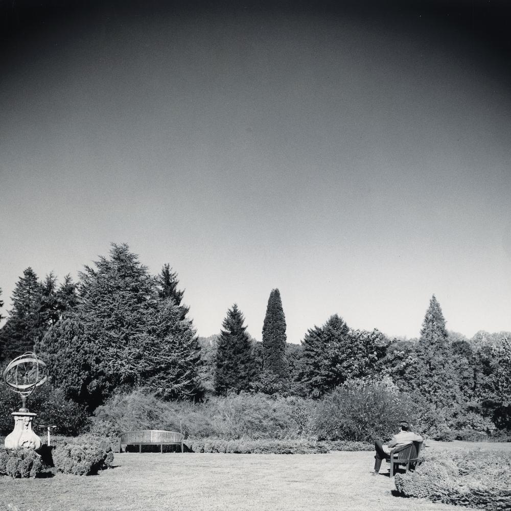 black and white photo of a large garden surrounding by trees, man sitting on bench