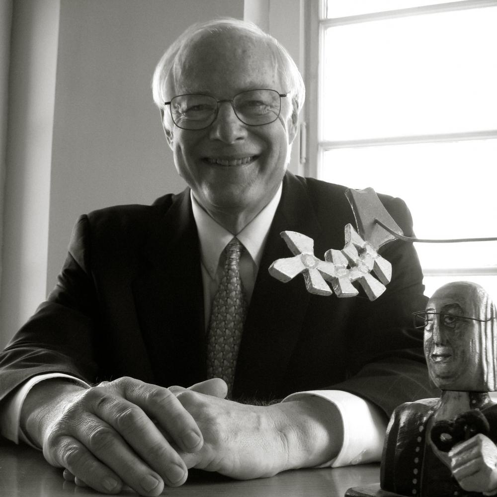 Chairman Leach smiles at the camera, sits with his hands folded on the table in front of him, wearing a dark suit and tie, and eyeglasses 