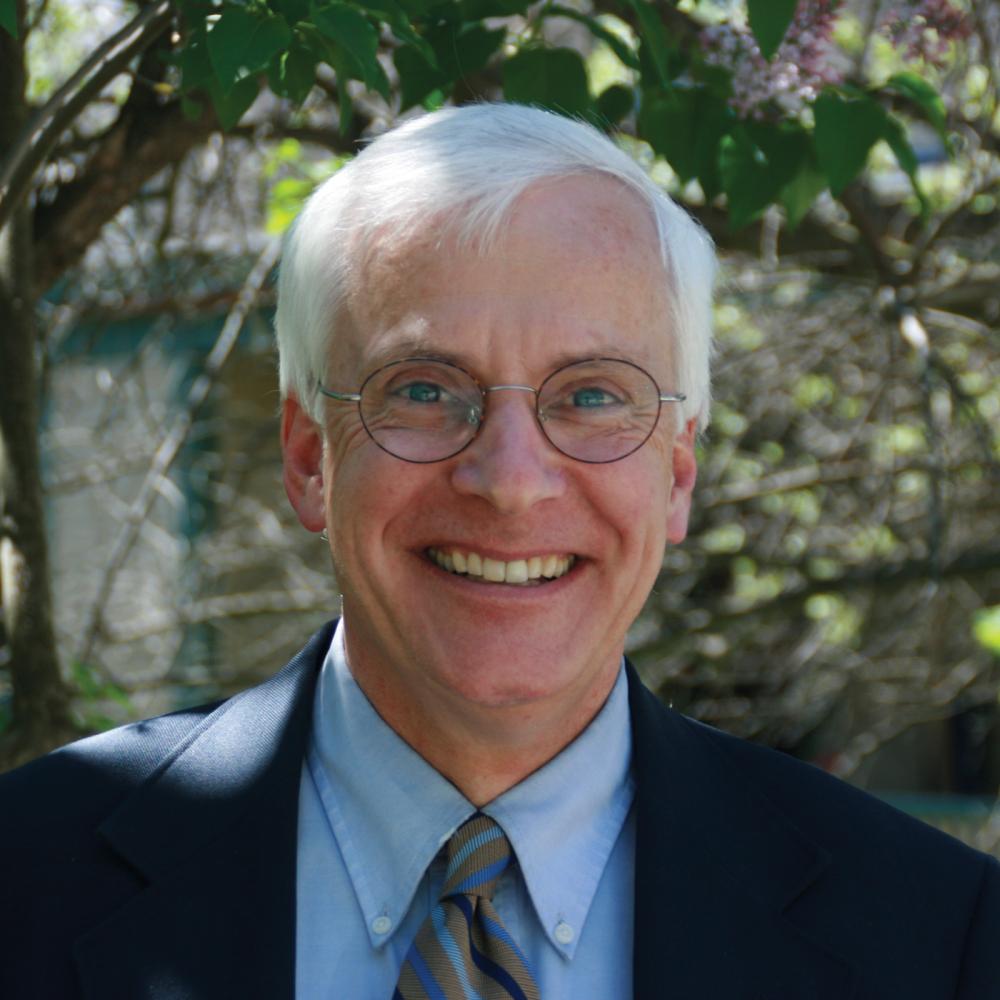 Gilbert smiles at the camera, wears glasses a blue suit and striped tie