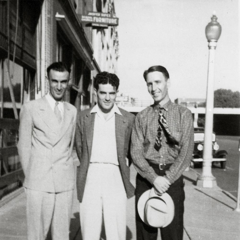 Three young men in light colored suits stand with their arms around each other; Thompson holds a straw hat