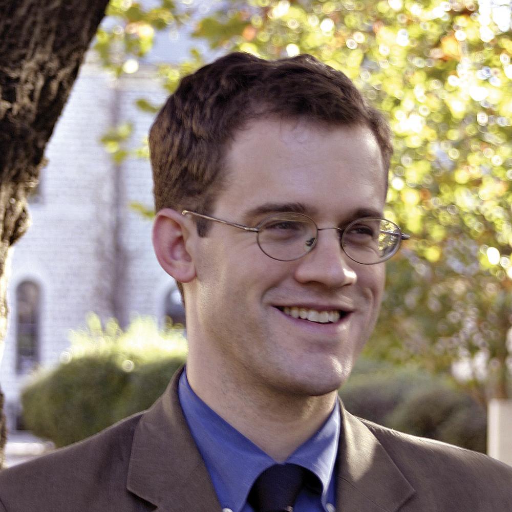 Nel, in a brown suit, blue shirt and eyeglasses, smiles; in the background is a white building and a leafy tree