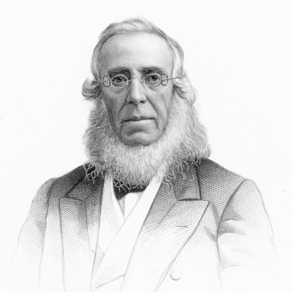 Cooper wearing octagonal eyeglasses, with a white beard no mustache, in a suit coat and bowtie