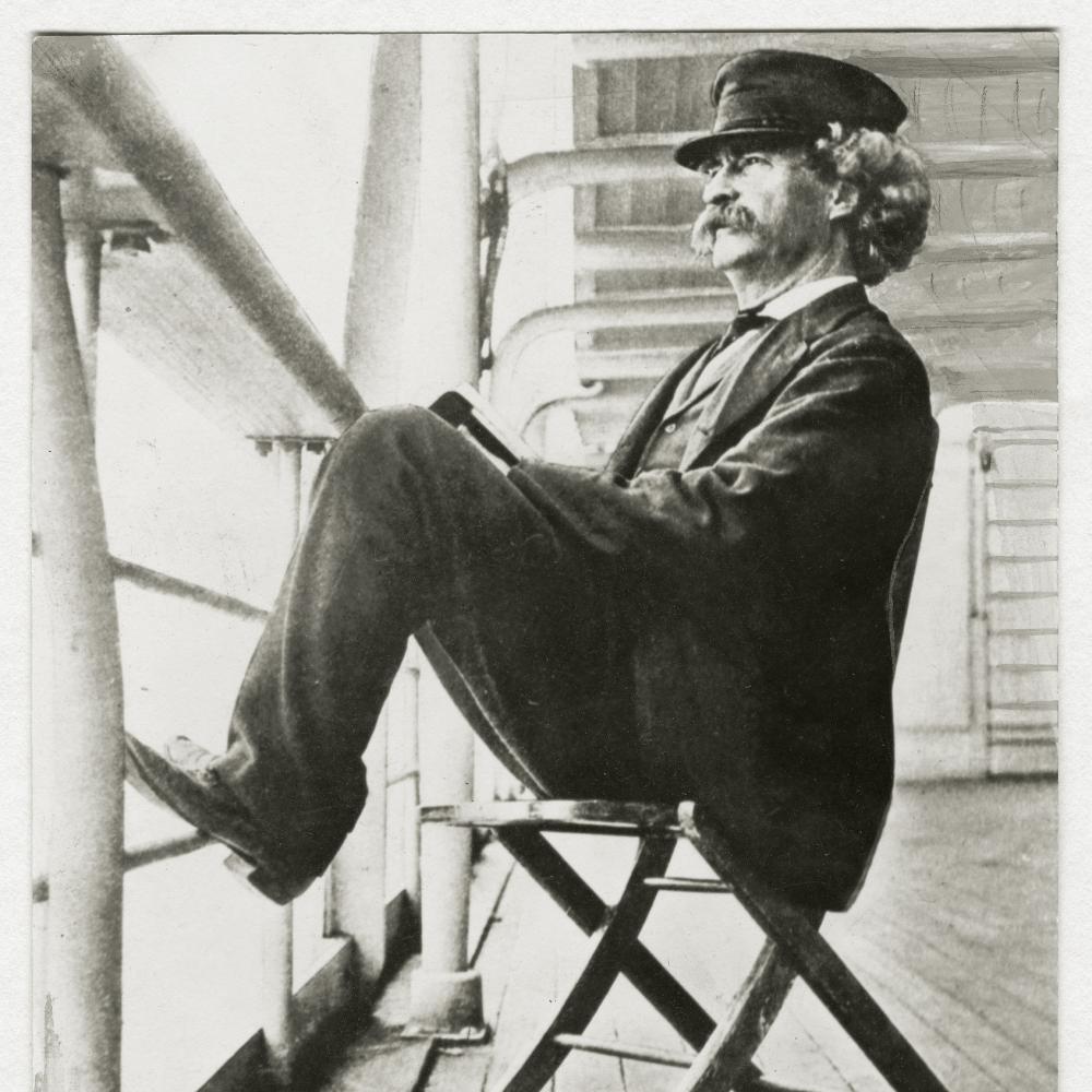 Black and white photo of Mark Twain sitting on a chair on the deck of a cruise ship.