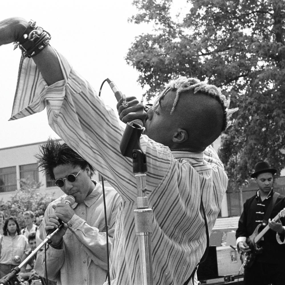 Black and white photo of an African American man performing on stage wearing a striped dress shirt.