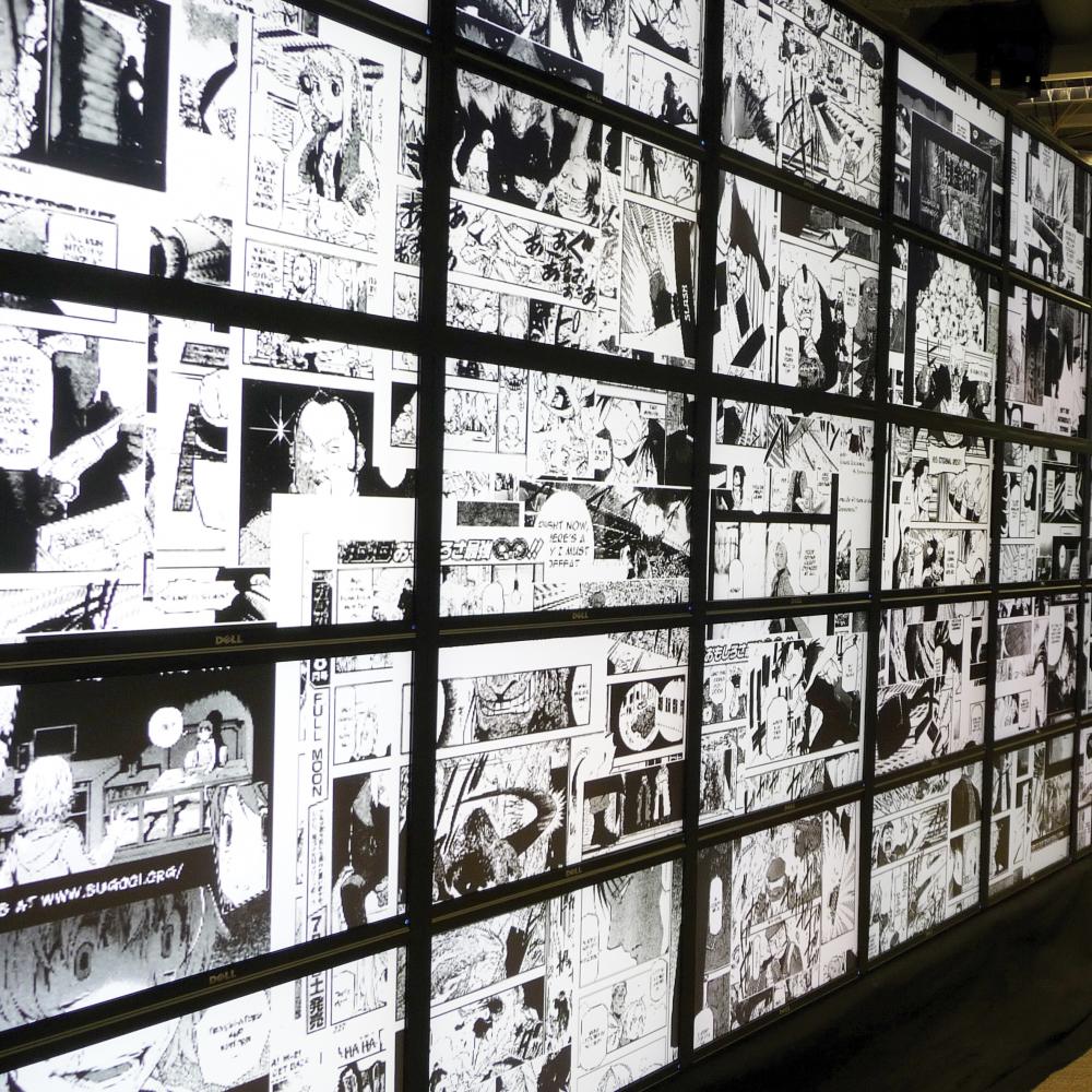 Two men stand in front of numerous small screens lit up with black and white manga illustrations