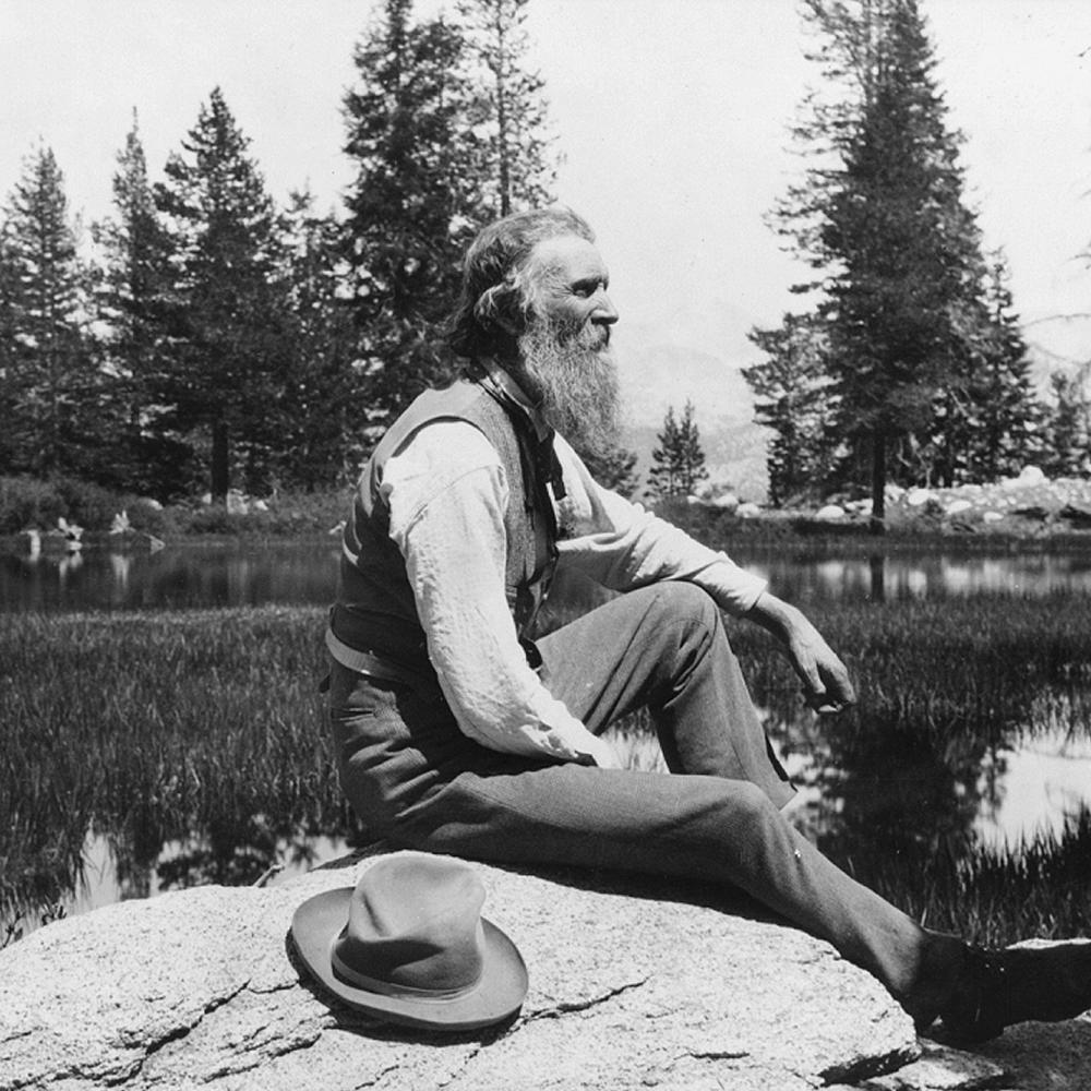 Muir sitting on a rock next to a lake, with a long gray beard, hat resting next to him