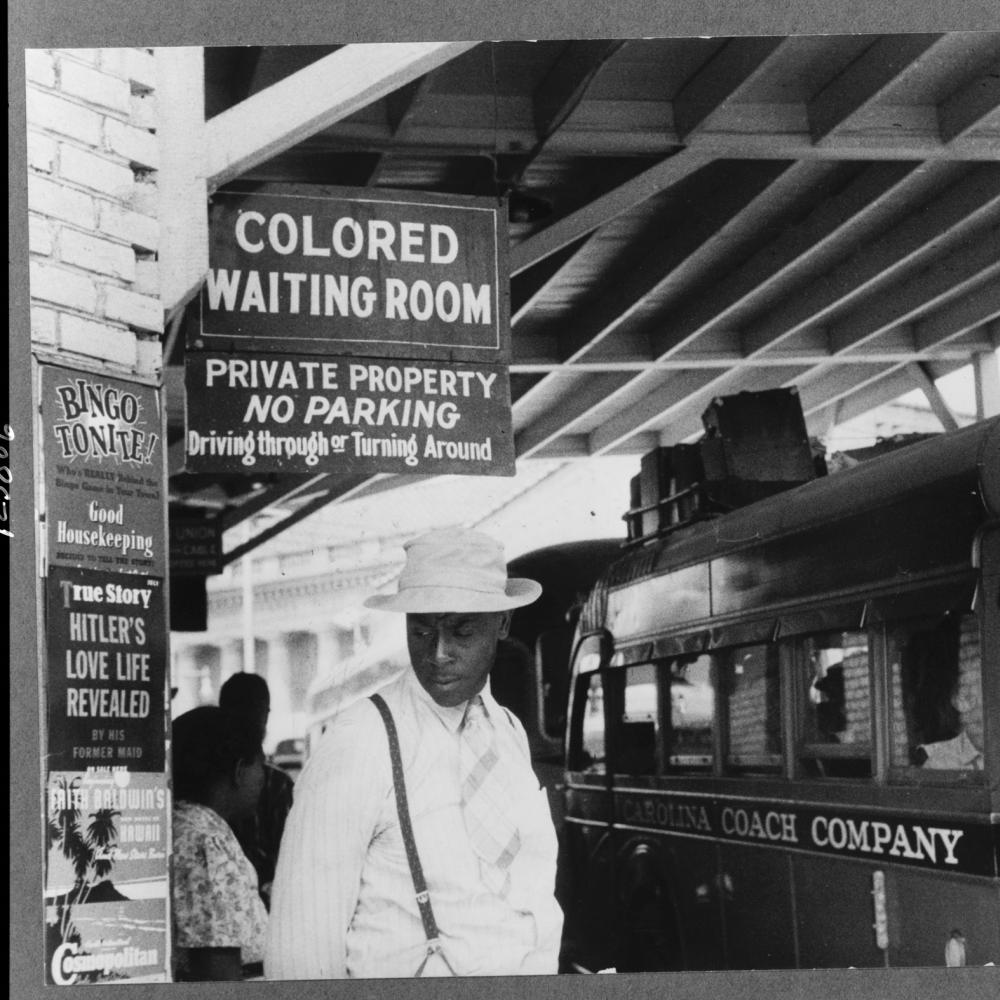 An African American man in a white hat heads toward the segregated waiting room while a bus idles at the curb
