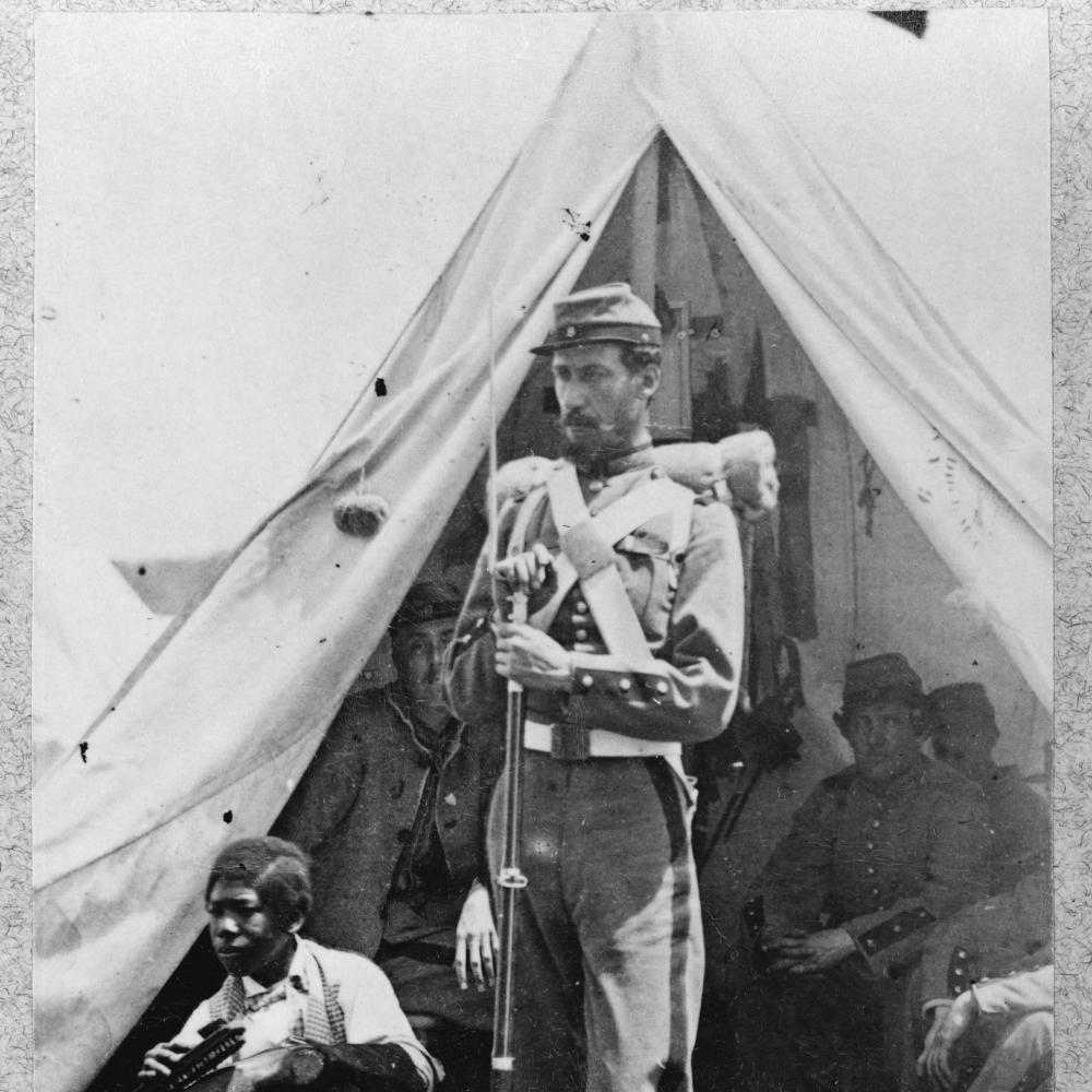 A soldier in full uniform, holding a bayonet, stands in front of a tent, where his fellow soldiers are sitting