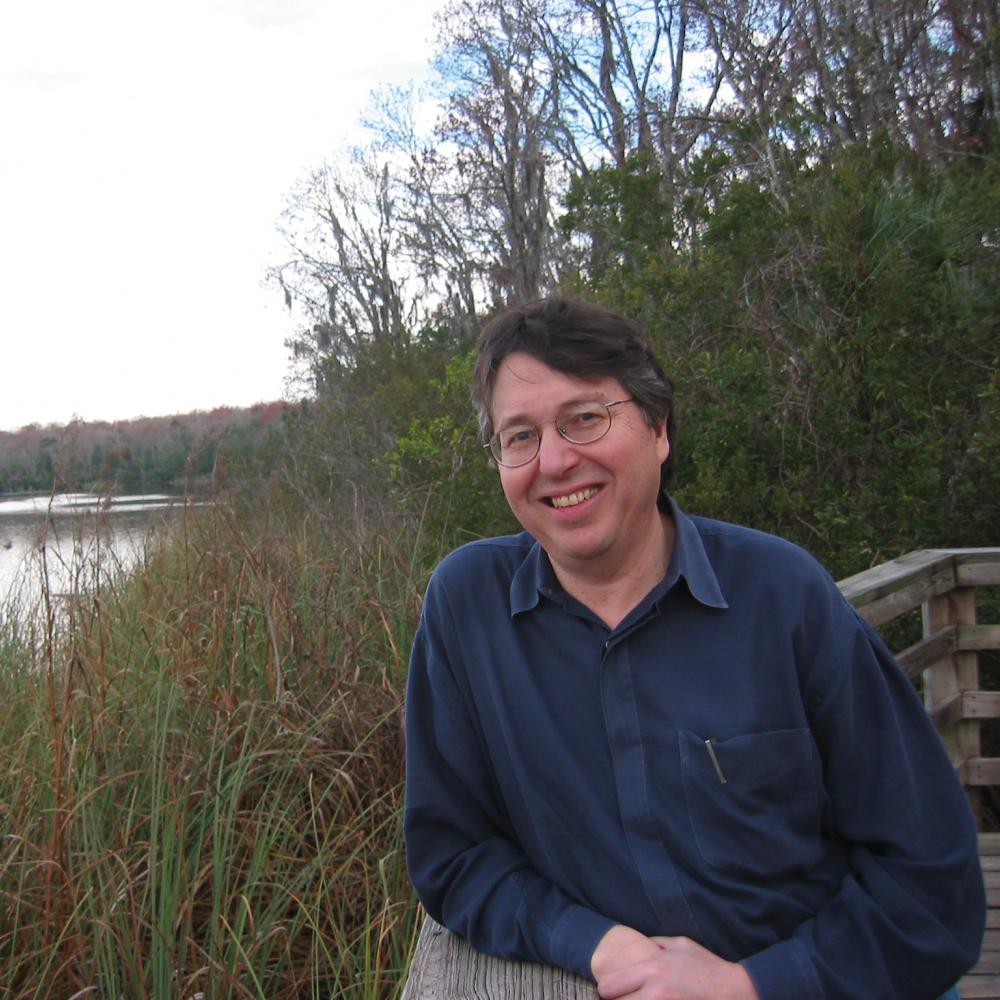 Hyde in a blue shirt, leaning on a railing overlooking a grassy pond