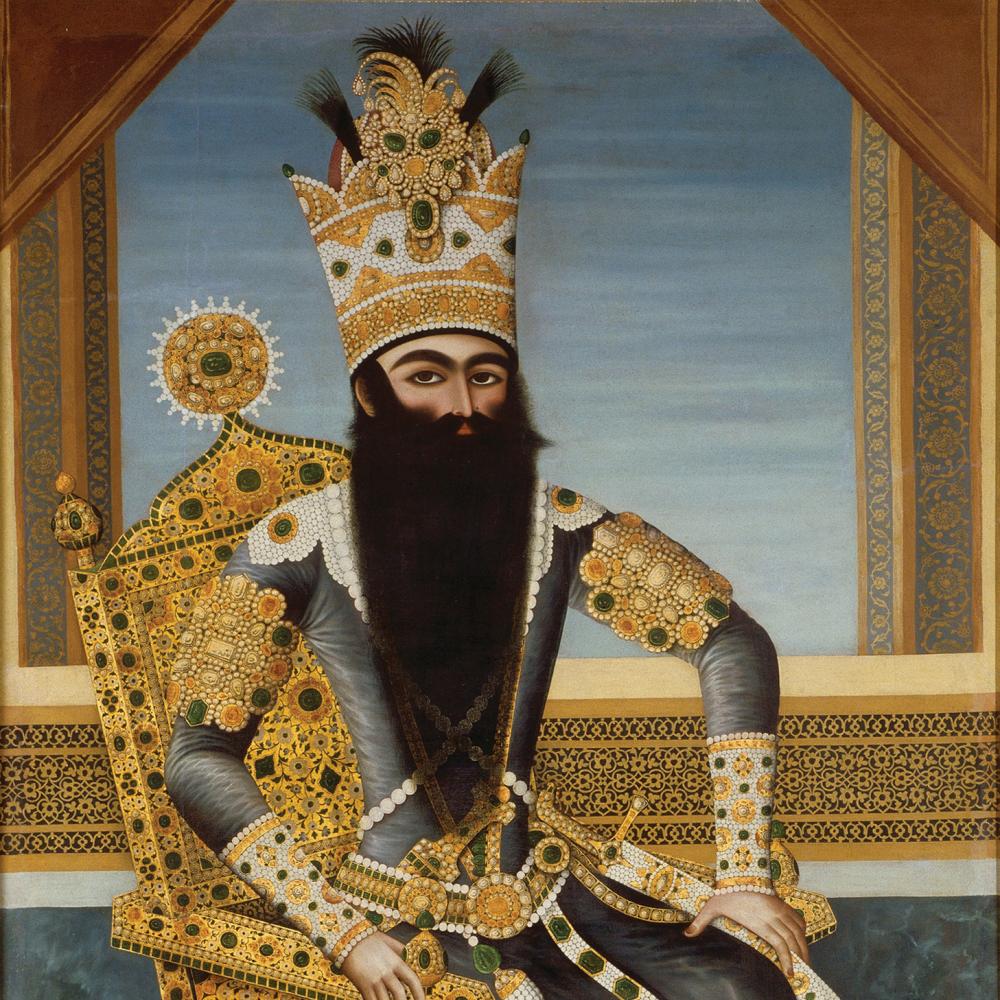 The Shah, with a long black beard, sitting on a richly decorated gold throne, wearing blue robes, gold jewelry, and a tall gold crown encrusted with gems